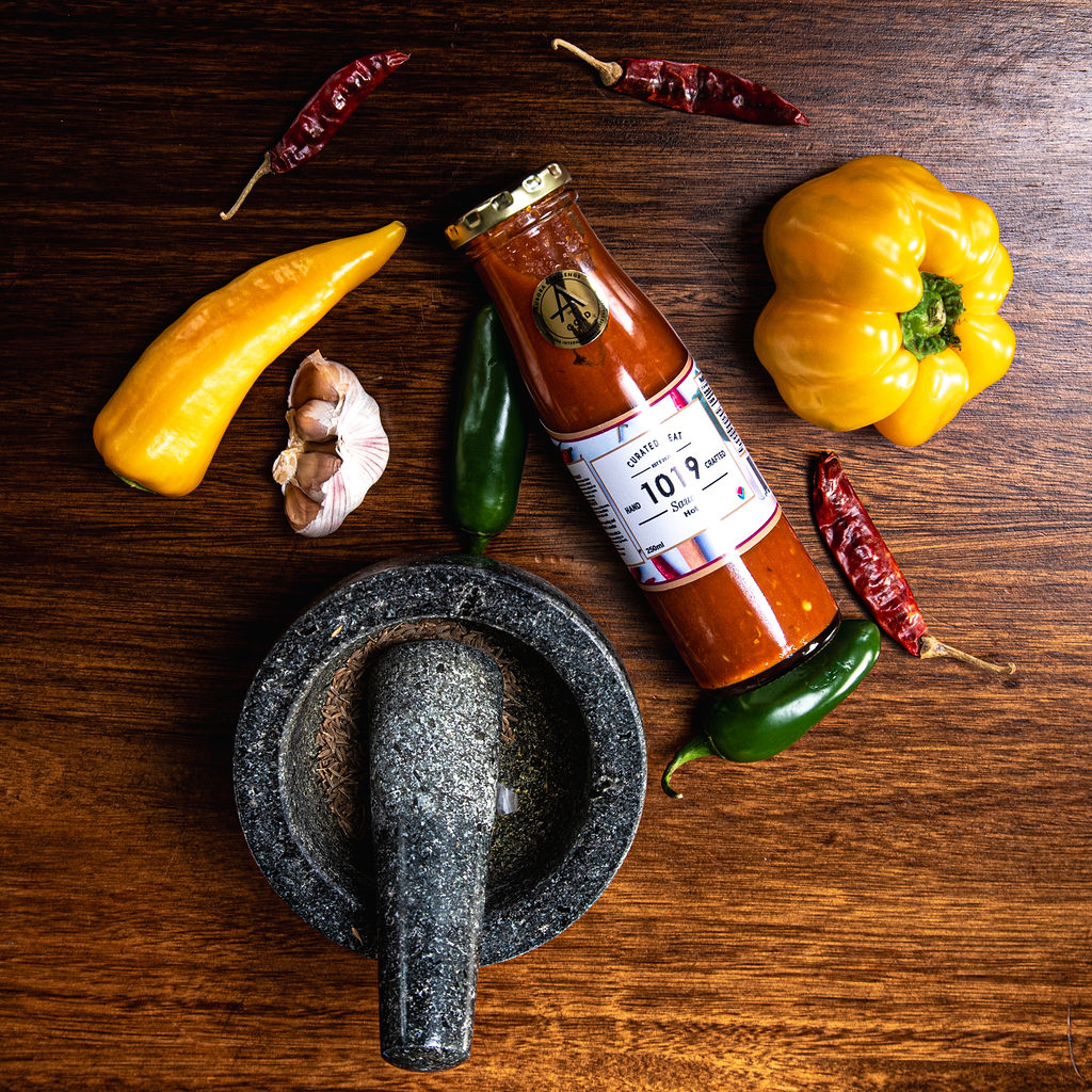 A bottle of 1019 Hot Sauce on a wooden surface, surrounded by a yellow bell pepper, yellow chili, green chili, red dried chilies, garlic, and a stone mortar and pestle.